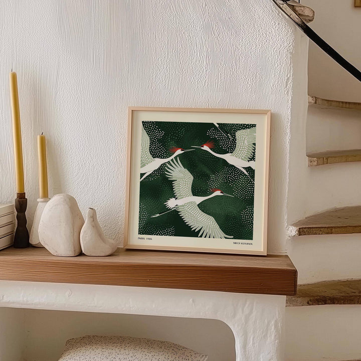 Print of White Crane Birds Art with green background leaning on a wooden shelf beside white ceramic ornaments