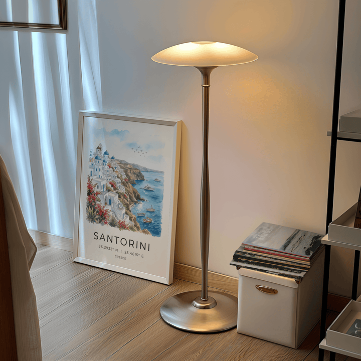 Print leaning on a floor of Santorini art print featuring cliffside village and blue sea with a floor lamp nearby