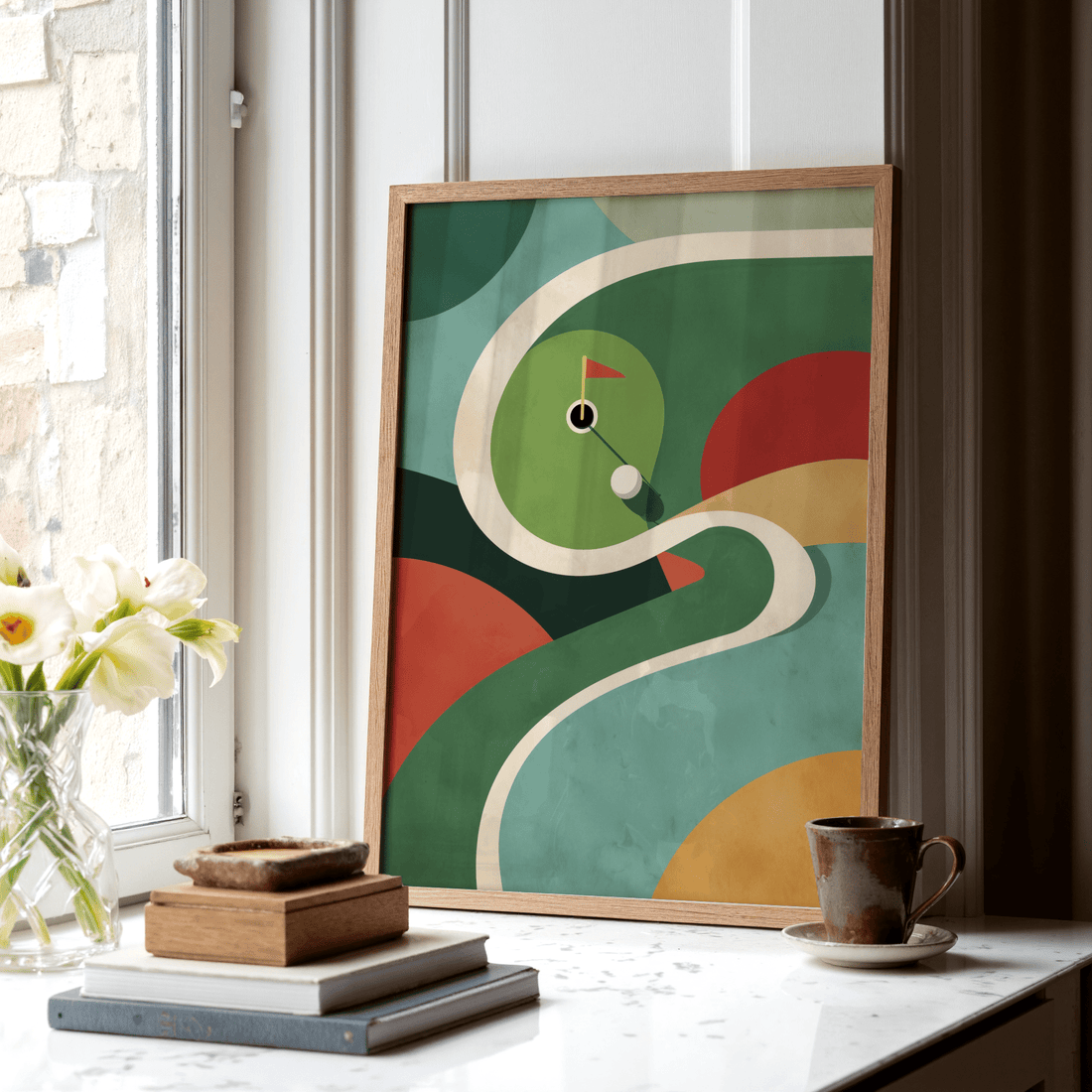 Colourful golf course print leaning in a sunny window beside white flowers, a stack of books and a mug