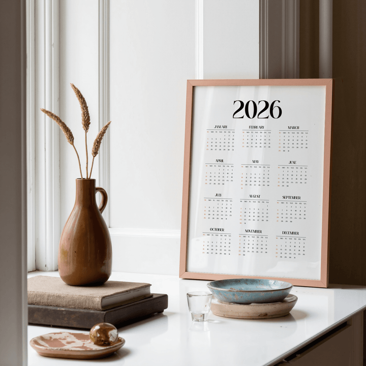 Minimalist wall calendar framed print with black and orange text on a white background leaning on a sideboard with pottery in a room