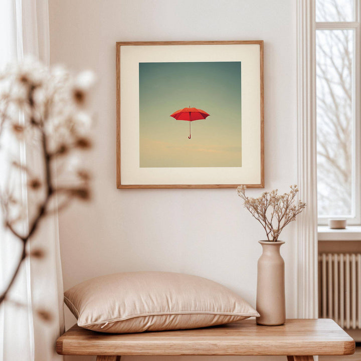 Surreal wall art of a red umbrella floating against a muted sky above a wooden bench with a cushion and vase