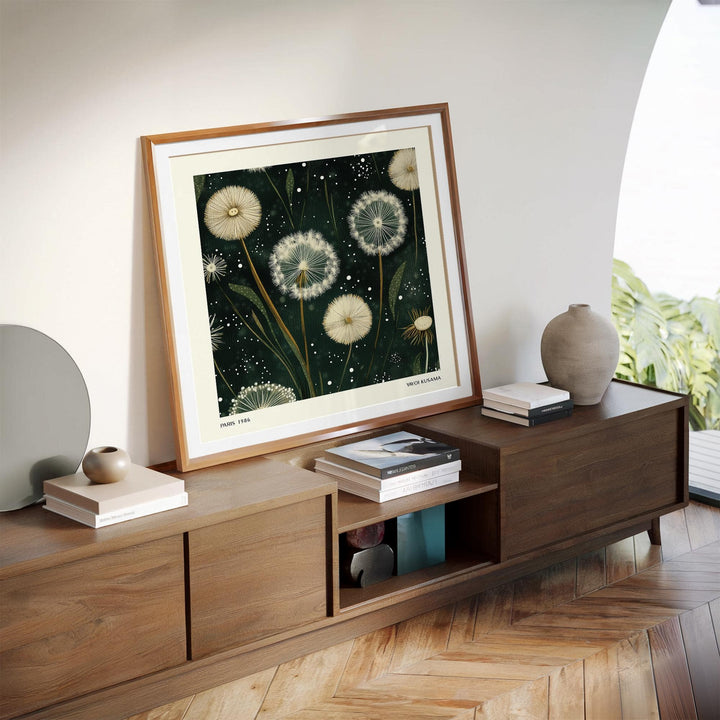 Square wall art of white dandelion seed heads leaning on a wooden sideboard with books and a vase