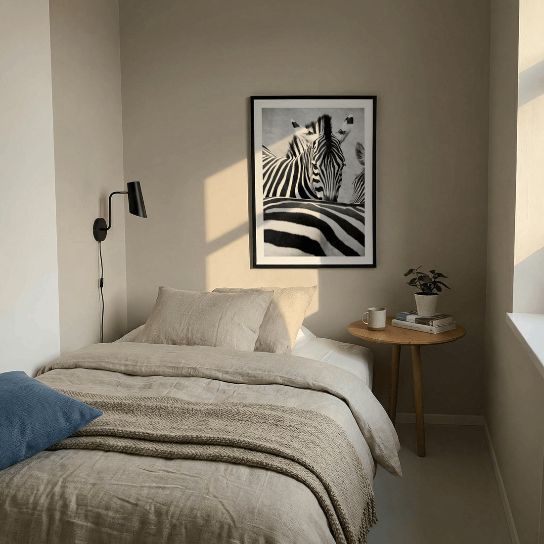 Small bedroom in soft evening light with linen bedding, a blue cushion, a black wall lamp and a black and white zebra print in a black frame