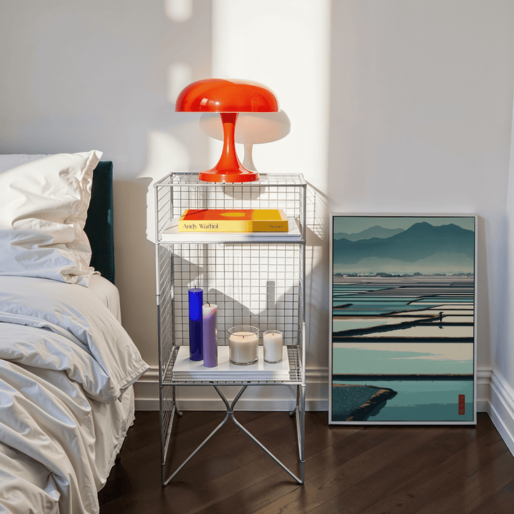 Print leaning on a floor showing serene rice field poster in minimalist style beside bedside table with orange lamp