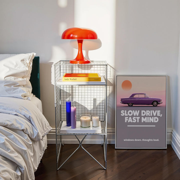 Vintage car wall art leaning on the floor next to a silver shelf and an orange lamp in a bedroom