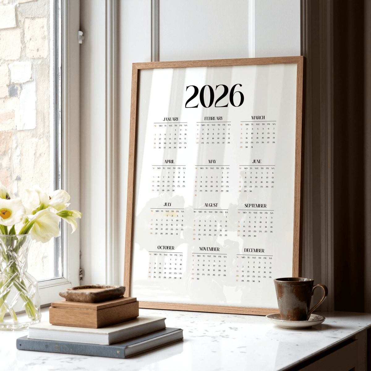 Minimalist wall calendar framed leaning on a window ledge with books and a cup nearby