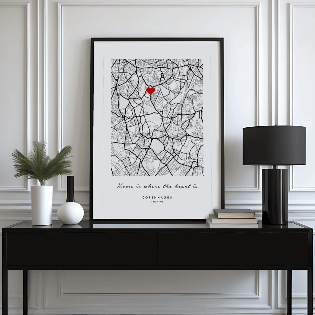 Black and white city map print with a small red heart, leaning on a black console table beside a lamp and books