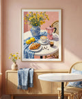 Framed artwork of a table setting with flowers and coffee on a pink wall.