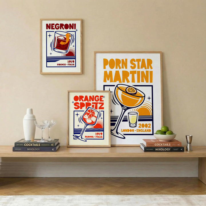 Three framed cocktail-themed art prints are displayed on a beige wall above a wooden shelf with bar decor. The prints feature retro illustrations: 'Negroni 1919 France - Italy', 'Orange Spitz 1919 Paris - France', and 'Porn Star Martini 2002 London - England'.