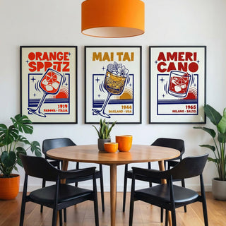 A styled dining room scene with a round wooden table, four black chairs, an orange pendant light, and three framed retro-style posters on the wall reading 'Orange Spitz', 'Mai Tai', and 'Ameri Cano'.