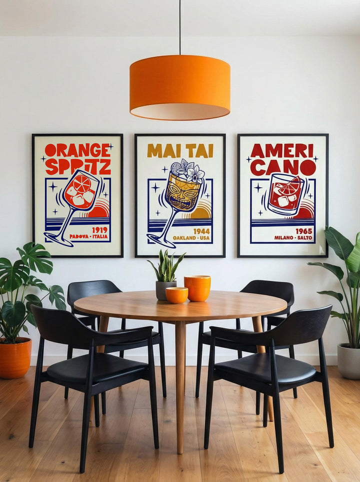 A styled dining room scene with a round wooden table, four black chairs, an orange pendant light, and three framed retro-style posters on the wall reading 'Orange Spitz', 'Mai Tai', and 'Ameri Cano'.