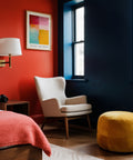 Modern bedroom with red wall, white chair, yellow ottoman, and abstract color block art.