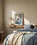 Bedroom with a bed, pillows, and a framed picture on a shelf with cat reading a book