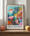 Framed Yayoi Kusama artwork on a wooden shelf with a vase and rolled fabric.
