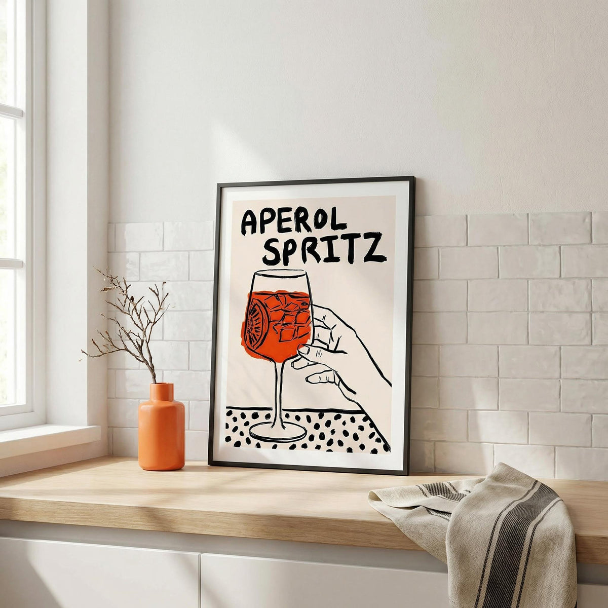 Framed artwork of a hand holding an Aperol Spritz glass on a wooden shelf.