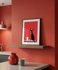 Framed artwork of a black cat on a red background in a room with a red wall and wooden shelf.