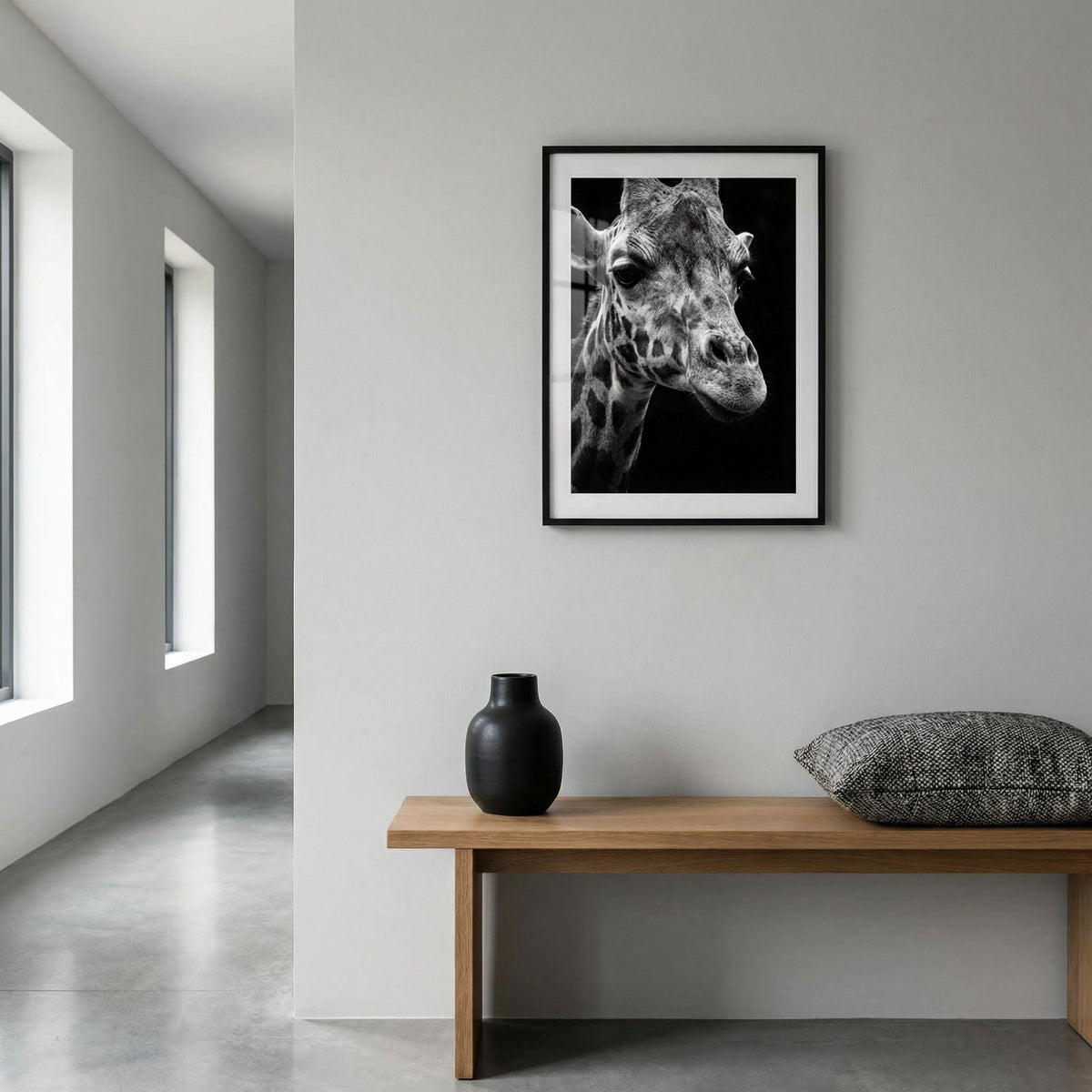 Framed black and white giraffe print on a wall above a wooden bench with a vase and pillow.
