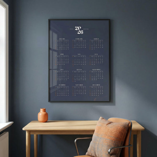 Framed calendar on a wall above a wooden desk with a chair and vase.