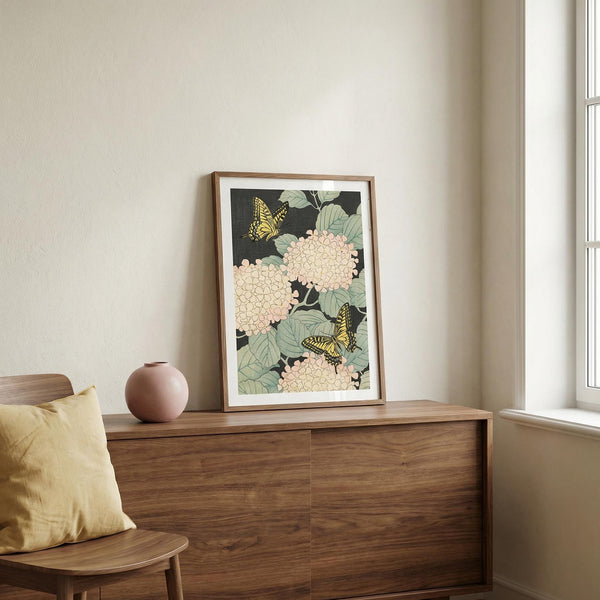 Framed floral and butterfly artwork on a wooden dresser with a window in the background.