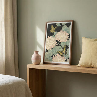 Framed floral artwork on a wooden side table with a vase and pillow against a neutral wall.