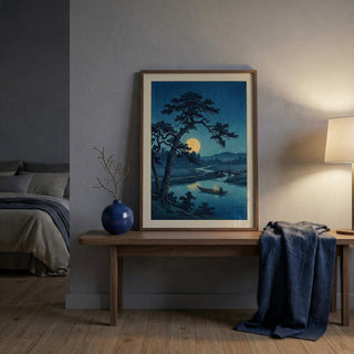 Framed artwork of a landscape with a tree and moon over a body of water in a room with a bed and lamp.