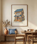 Framed artwork of a building on a wall above a wooden table and chairs in a room with natural light.