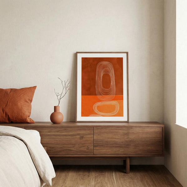 Framed abstract art on a wooden dresser with a pillow and vase in a bedroom setting.