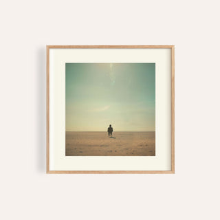 Framed print of minimalist surrealism art with a person sitting in a pale desert under a pale blue sky