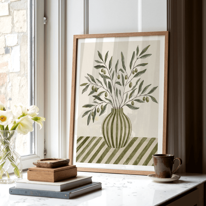 Framed Olive Branch Art print with green and brown hues leaning on a window sill beside a cup and books