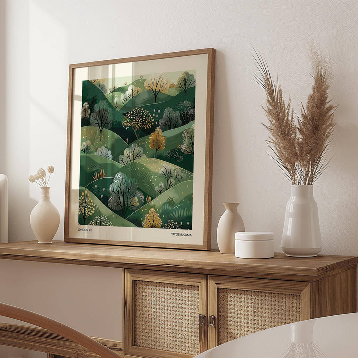Large nature wall art of green hills with trees in an illustration style leaning on a wooden sideboard beside neutral vases