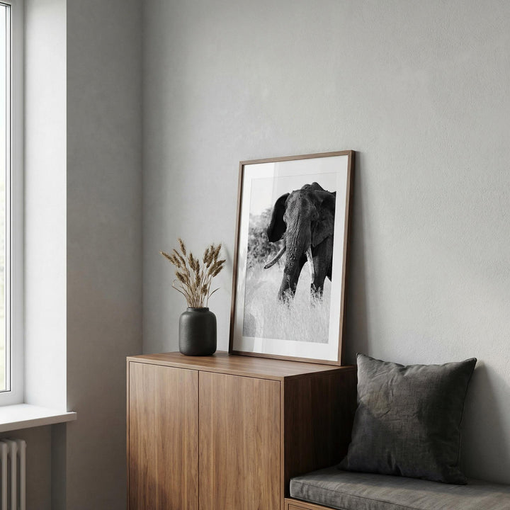 Elephant Black And White Art Print leaning on a wooden cabinet with a dark vase and dried flowers in a grey room