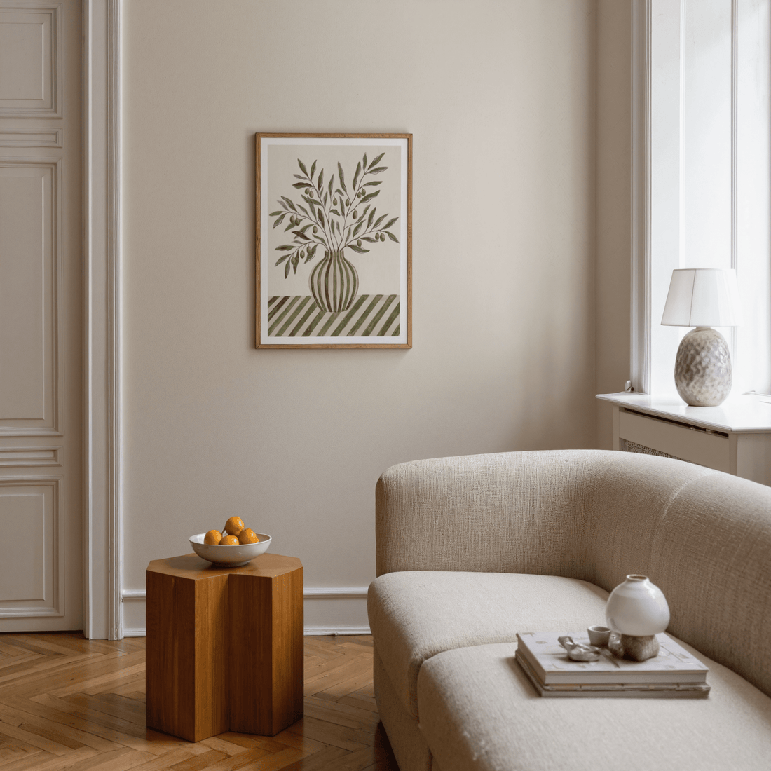 Olive branch print in soft green and sand tones on a warm beige wall above a cream sofa and a wooden side table