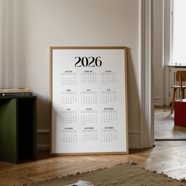 Minimalist wall calendar leaning on wooden floor near a green side table with books