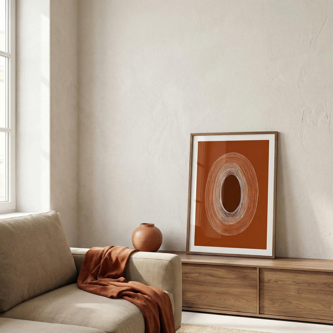 Neutral living room with a textured plaster wall, a beige sofa with a rust throw, a terracotta vase and a rust abstract print leaning on an oak sideboard