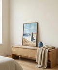 Wooden side table with a framed beach scene painting and a blue vase in a bedroom.