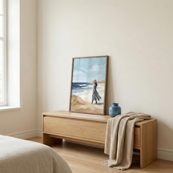 Wooden side table with a framed beach scene painting and a blue vase in a bedroom.