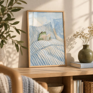 Frog Reading In Bed Print, a small A4 framed print leaning on a pale oak bookshelf in a living room