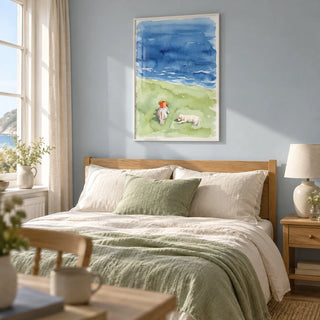 Reading By The Sea Art Print, framed on the wall of a restful bedroom in a UK coastal home with pale blue walls and natural linen curtains