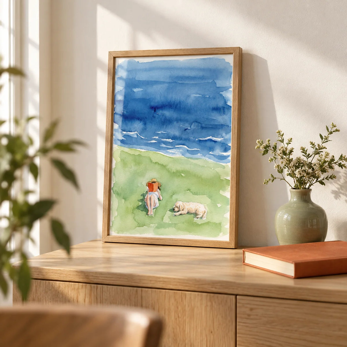 Reading By The Sea Art Print, a small A4 framed print leaning on a pale oak sideboard in a living room