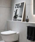 Une œuvre d'art humoristique dans la salle de bain avec l'inscription « Take a Seat » (Prenez place) au-dessus de toilettes modernes.